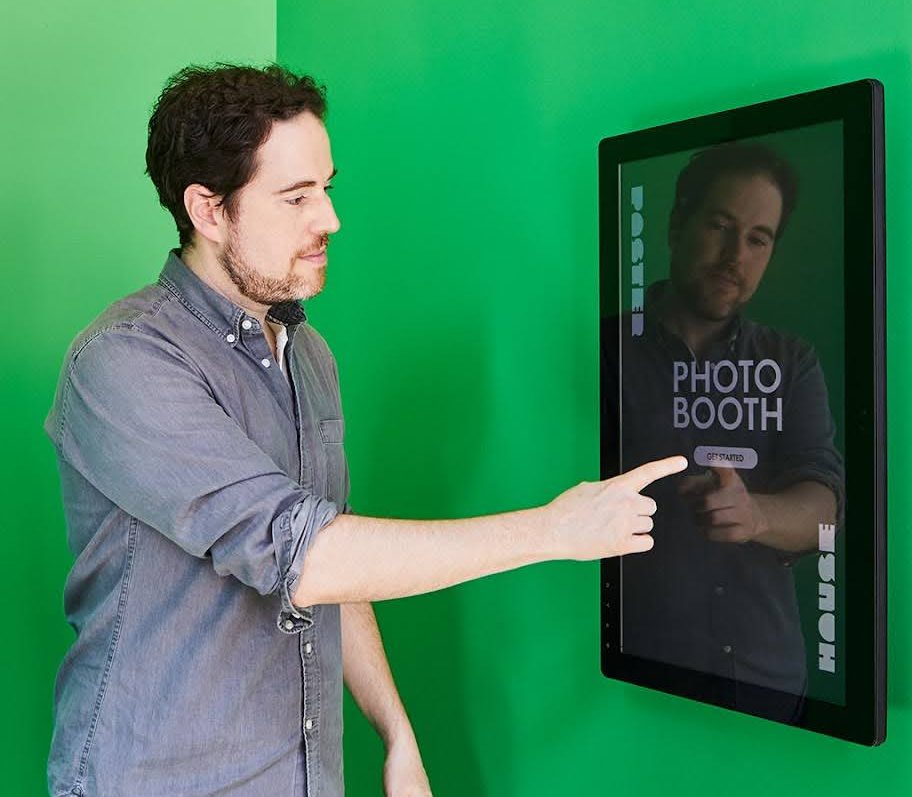 A figure touching a touch screen stating 'Photo Booth,' in a green room.