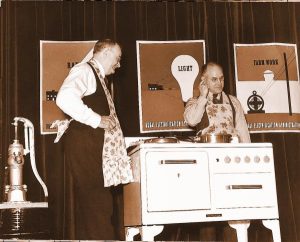 A photograph of 2 men showing off appliances in front of posters.