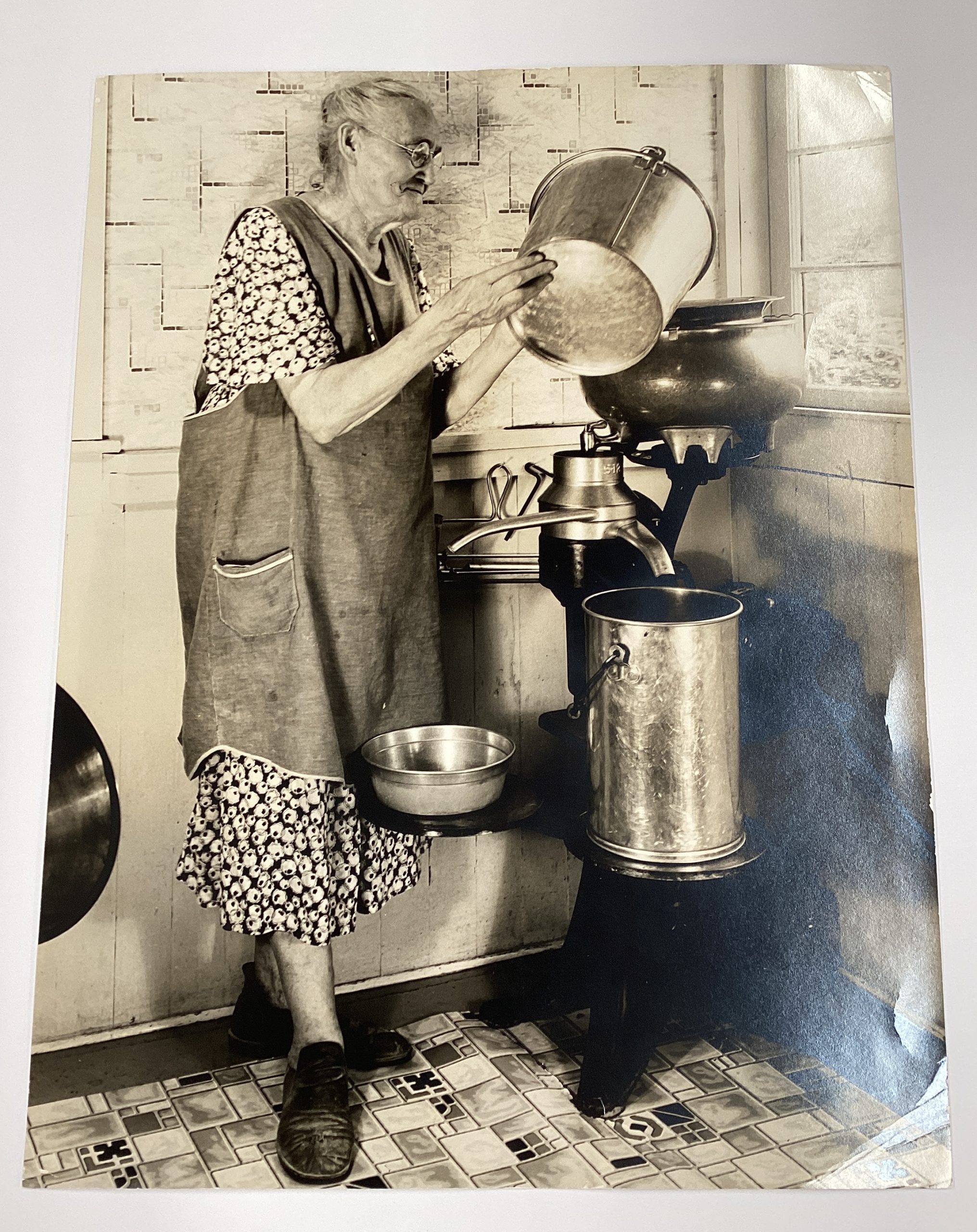 A black-and-white photograph of an older woman in an apron pouring liquid into an electric milk separator.