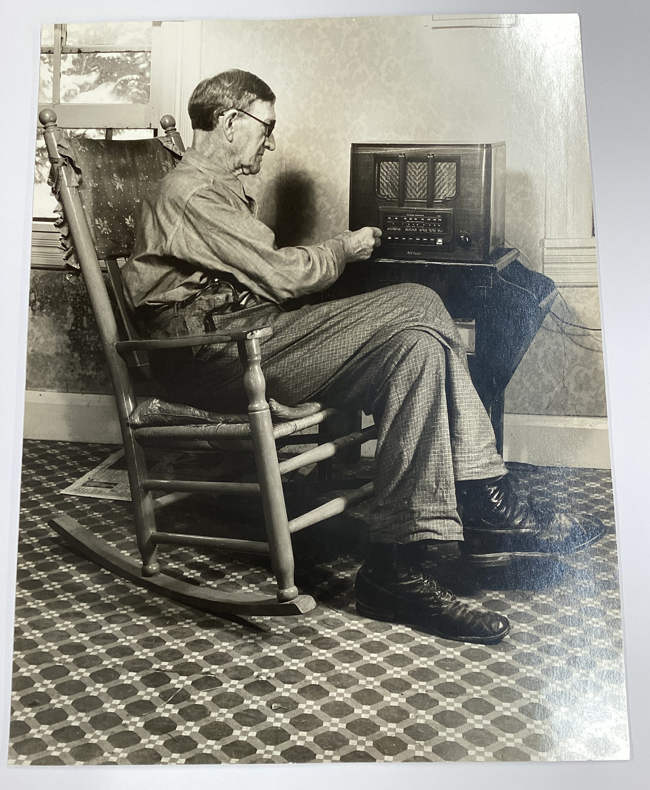 A black-and-white photograph of an older white man in a rocking chair turning the knobs on a radio.