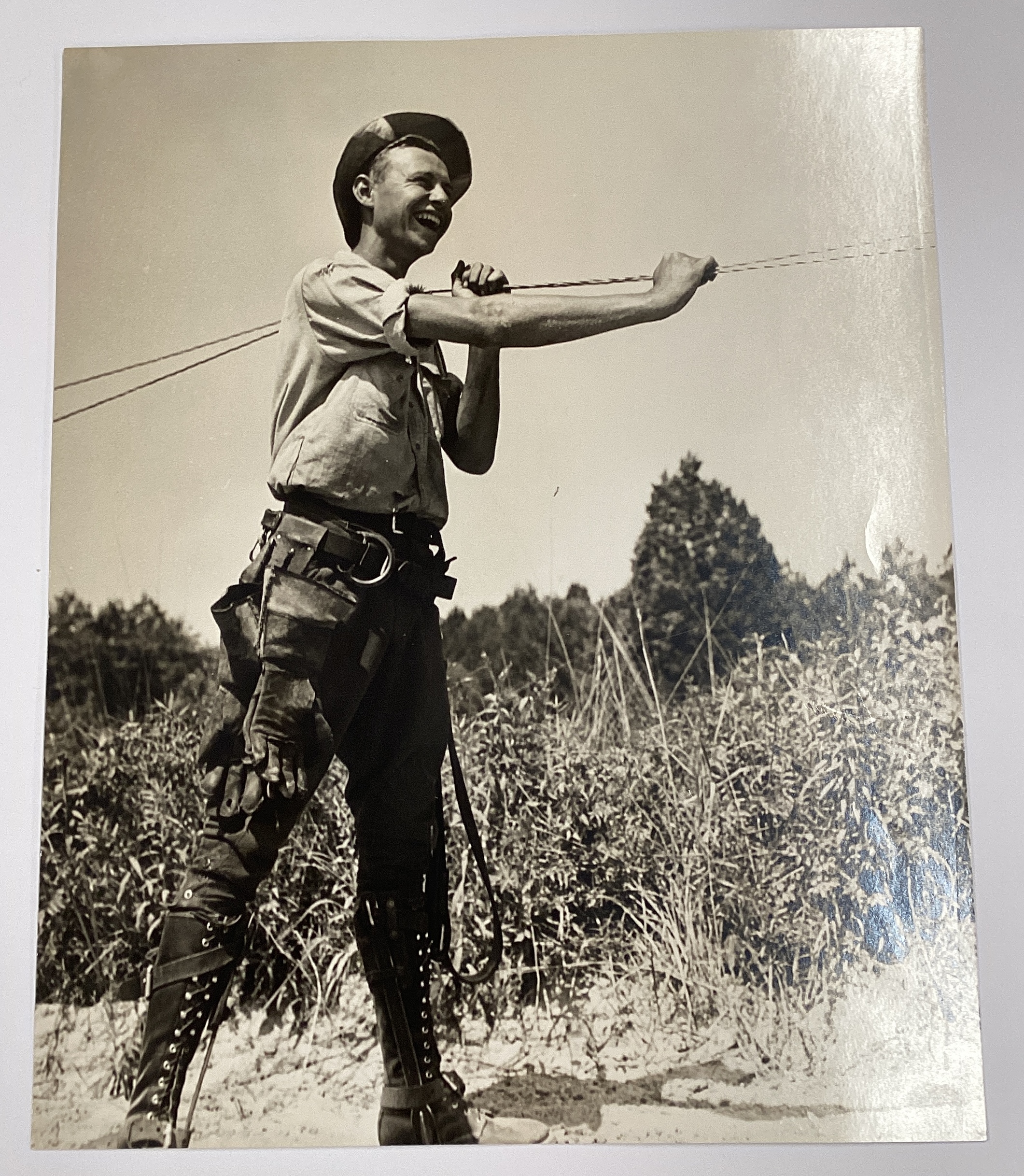 A black-and-white photograph of a smiling white electrical worker holding a power line in a field.