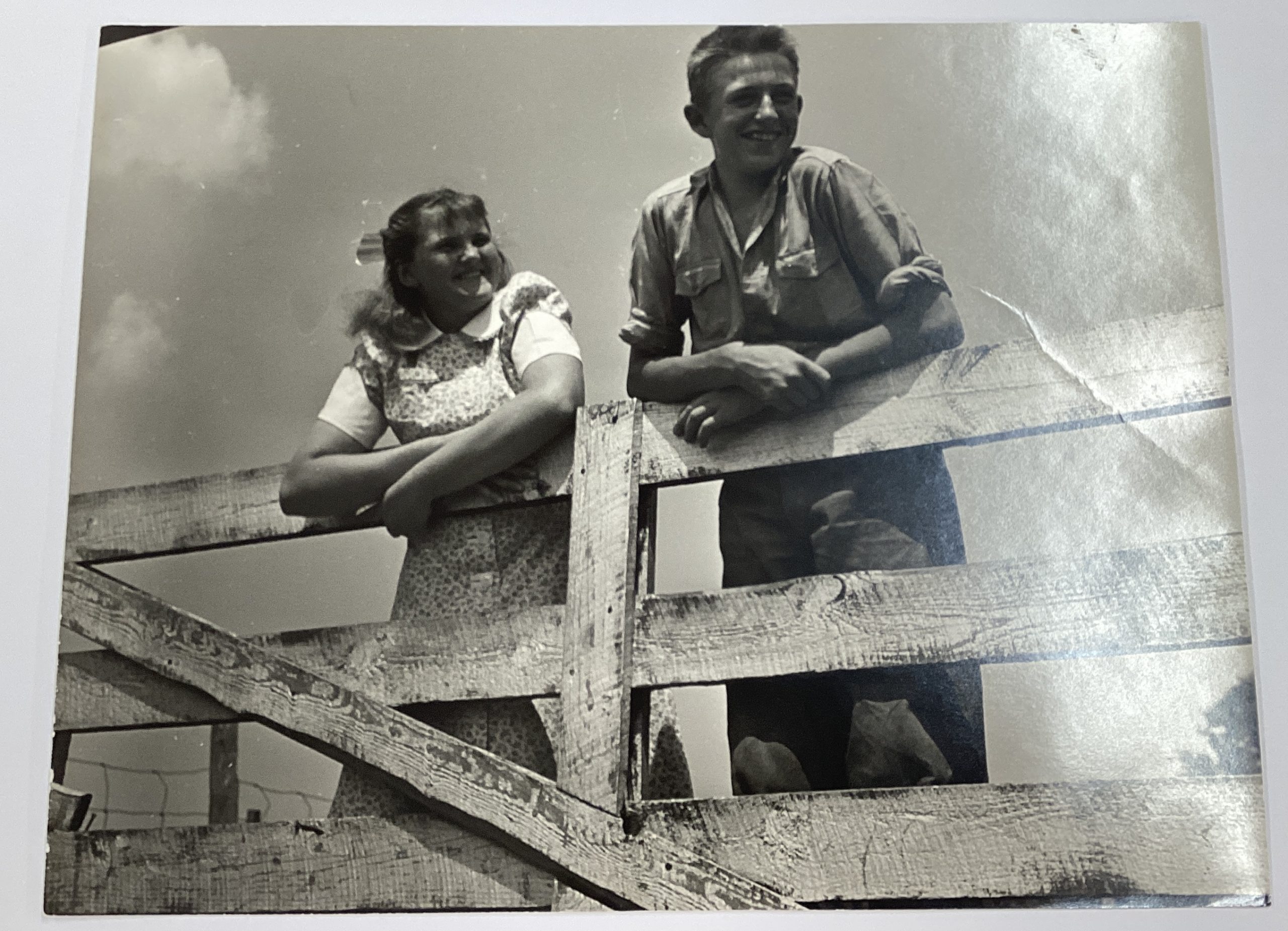 A black-and-white photograph of a smiling girl and boy, both white, hanging on a wooden gate.