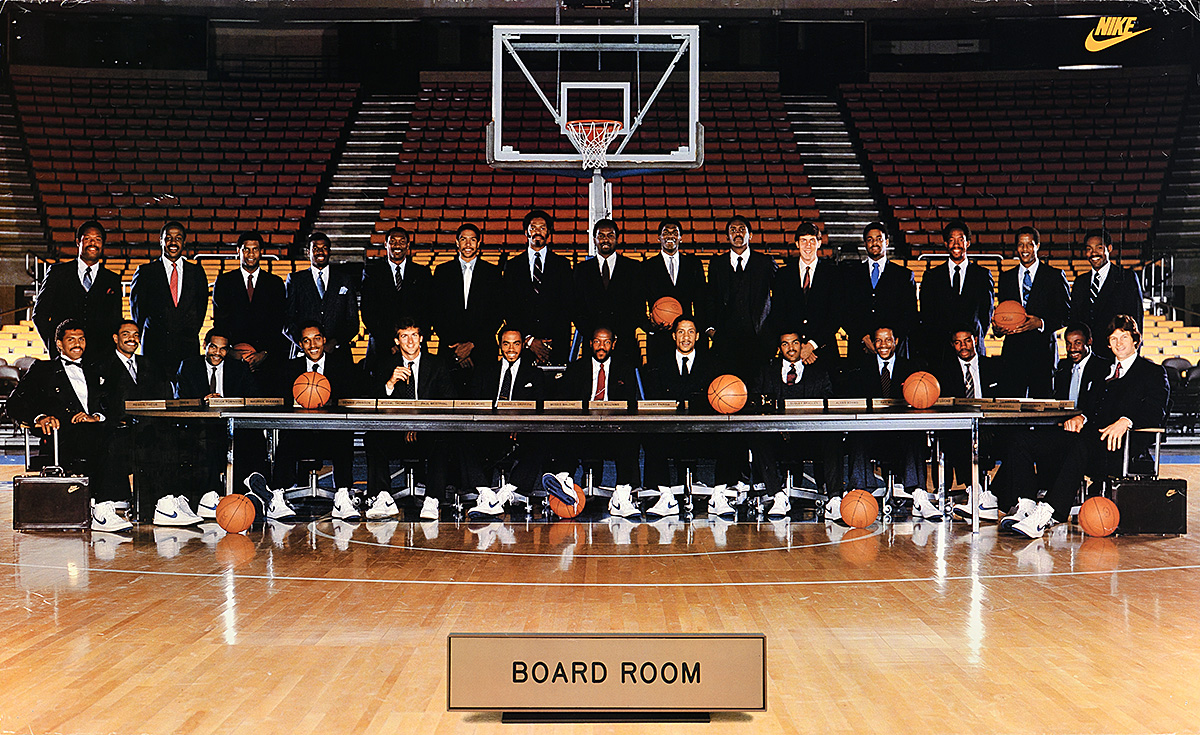 A poster of a large group of men in suits and white Nike shoes at a long table on a basketball court.