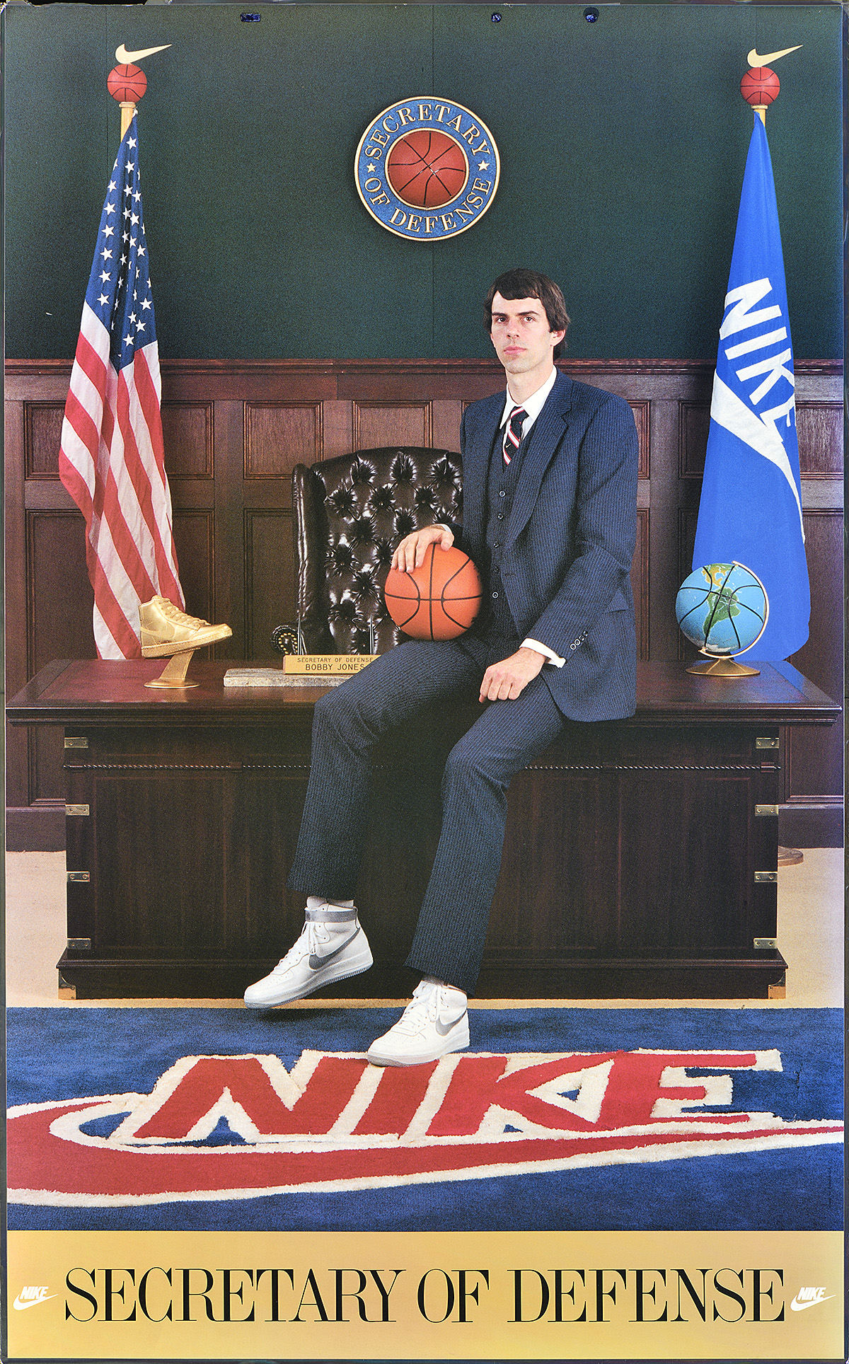 A poster of a tall white man in a suit sitting on an executive desk holding a basetball.