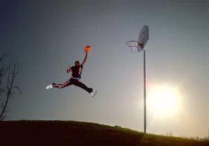 A photo of a man dunking a basketball while leaping against a setting sun.