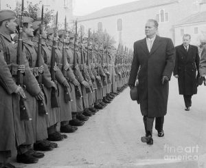 A black and white photograph of a stern white man walking past a row of guards with guns.