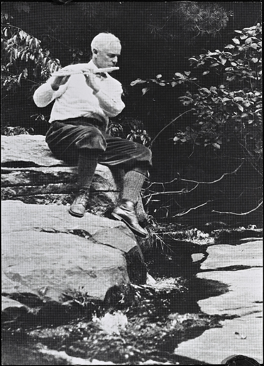 A black and white photograph of a white man playing a flute above a creek.