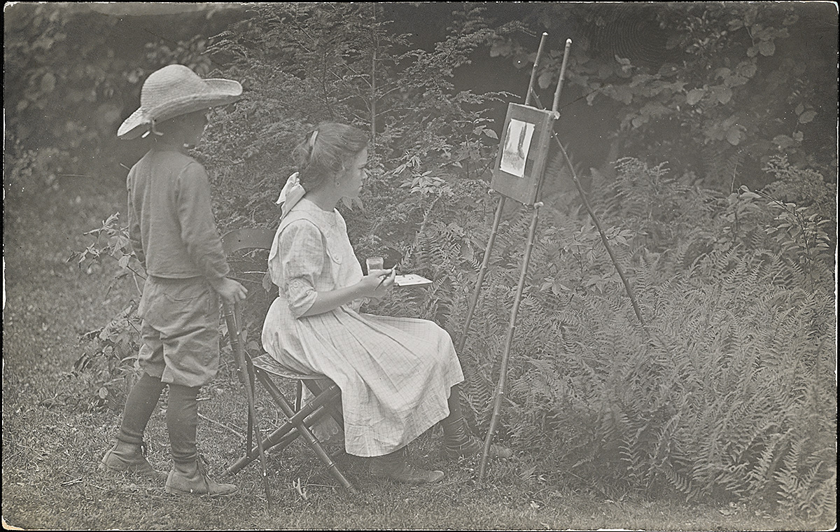 A black and white photograph of a white girl painting at an easel outdoors while a white boy watches.