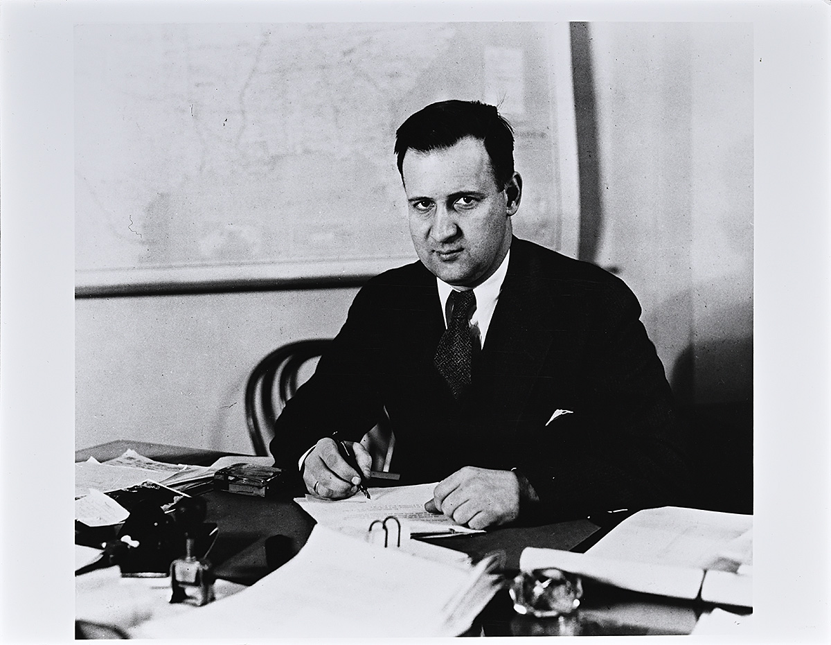 A black and white photograph of a white man in a suit signing papers at a desk.